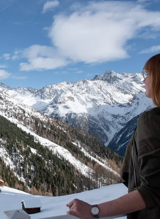 Eine Person steht auf einem verschneiten Balkon und blickt auf schneebedeckte Berge unter einem teilweise bewölkten Himmel.