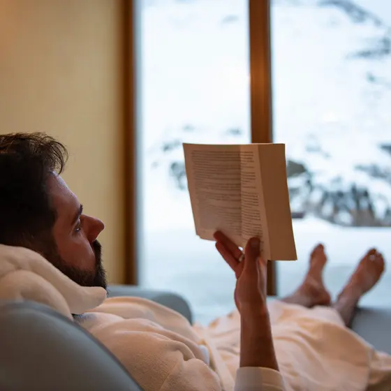 Ein Mann im Bademantel entspannt sich auf einer Couch und liest ein Buch, während draußen vor dem Fenster eine Schneelandschaft zu sehen ist.