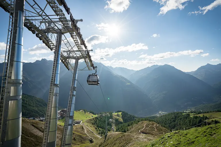 Eine Seilbahn, die einen Berg hinauffährt, mit Sonnenlicht über grünen Hügeln und fernen Berggipfeln im Hintergrund.