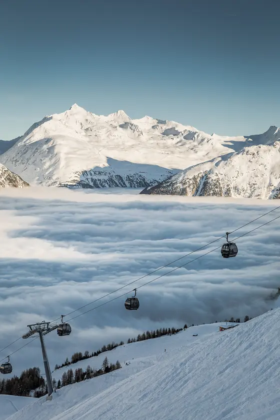 Bild einer weißen Piste im Skigebiet, während eine dicke Wolkenschicht das Tal bedeckt. Man sieht die fahrende Bergbahn und weiße Gipfel mit blauem Himmel im Hintergrund.