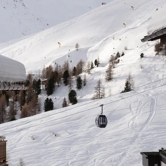 Ein Skilift fährt einen verschneiten, von Bäumen gesäumten Berghang hinauf, auf dem sich unter einem wolkenverhangenen Himmel zahlreiche Skifahrer tummeln.