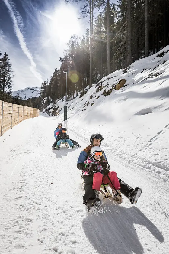 Bild einer Rodelbahn, auf der Erwachsene und Kinder auf Rodeln hinunterfahren. Links sieht man die Holzbande und rechts den schneebedeckten Wald.