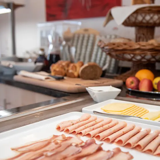 Schinken- und Käsescheiben auf einer Platte mit Brot, Obst und Gebäck im Hintergrund auf einem Frühstücksbuffet.