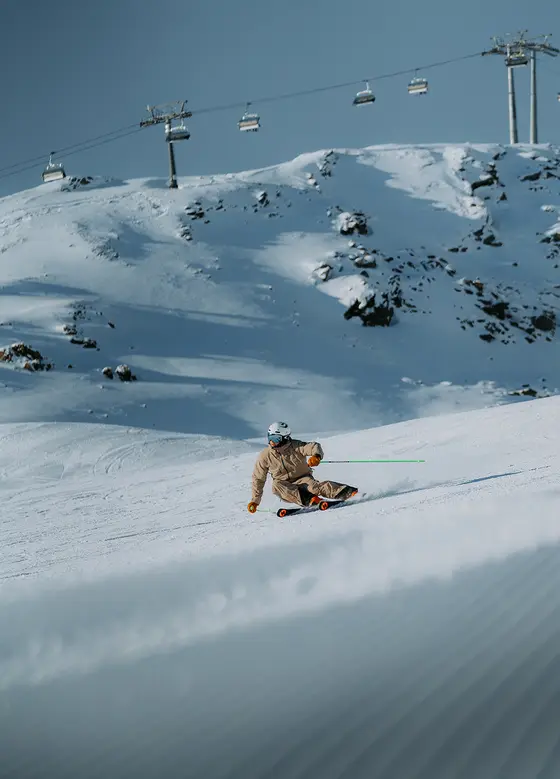 Eine Person fährt mit dem Snowboard eine verschneite Piste hinunter, mit Skiliften und Bergen im Hintergrund.