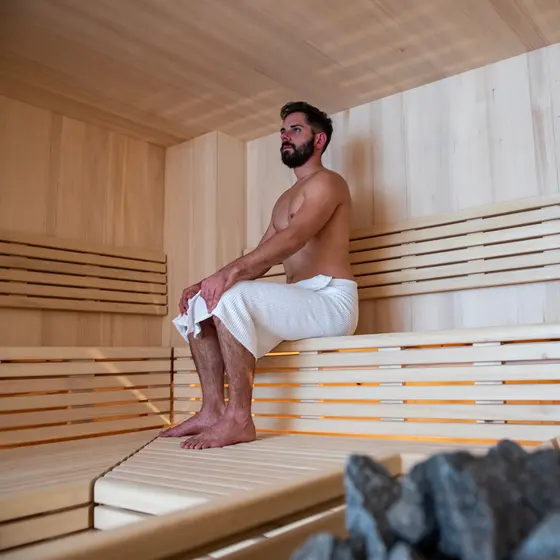 Ein Mann sitzt mit einem Handtuch auf einer Holzbank in einer Sauna, in der im Vordergrund heiße Steine zu sehen sind.