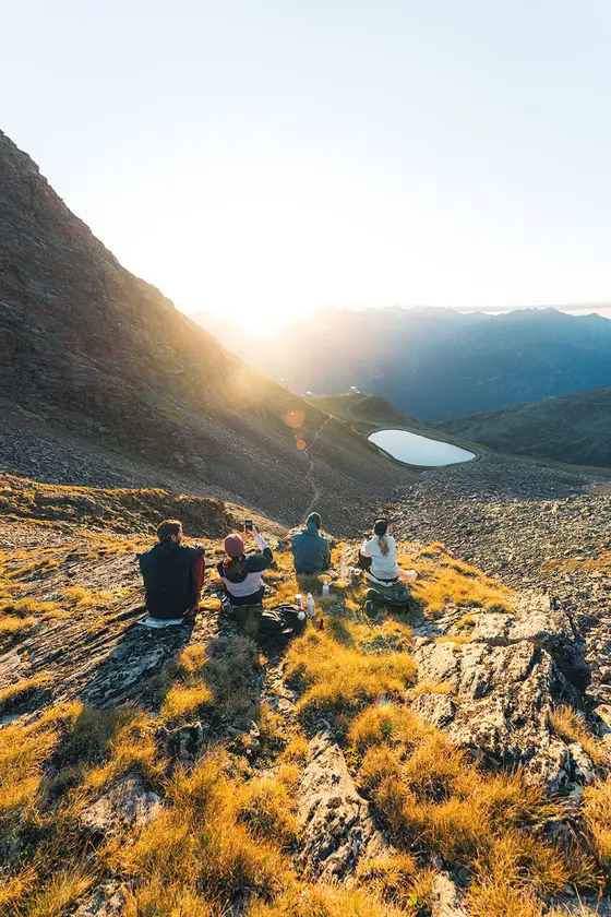 Mehrere Wanderer sitzen bei Sonnenaufgang auf einem grasbewachsenen Berghang und blicken auf ein Tal mit Seen und fernen Gipfeln.