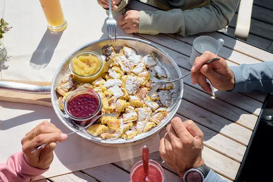 Ein Teller mit Kaiserschmarren steht in der Mitte eines Tisches und man sieht die Hände von drei Personen, die um den Tisch sitzen.