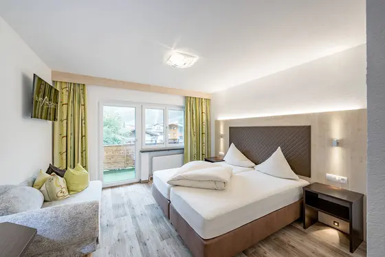 Helles, modernes Hotelzimmer mit Doppelbett, Sofa, grünen Vorhängen und einem Balkon mit Sitzgelegenheiten im Freien.