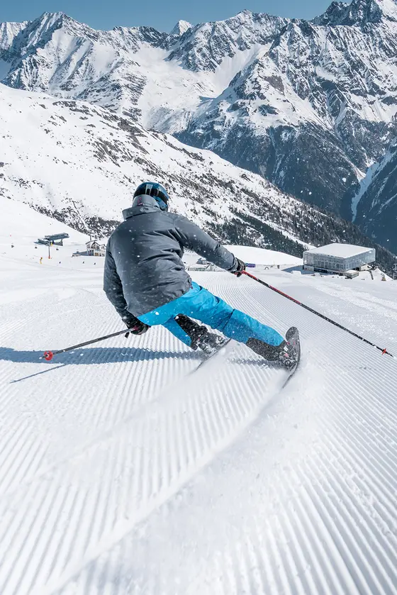 Ein Skifahrer in blauer Hose und grauer Jacke fährt eine präparierte Schneepiste mit Bergen im Hintergrund hinunter.