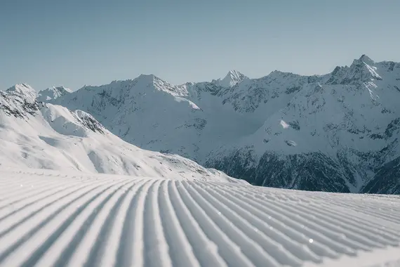 Präparierte Skipiste mit parallelen Linien im Vordergrund und schneebedeckten Bergen im Hintergrund.
