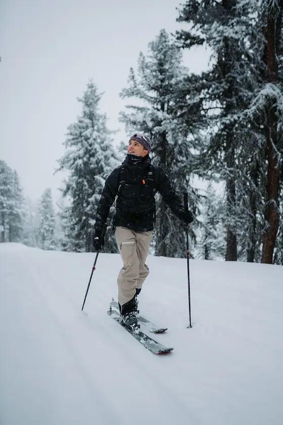 Zwei Skifahrer in farbenfroher Ausrüstung wandern auf einem verschneiten Berg bergauf und tragen dabei Skier auf ihren Rucksäcken unter einem klaren blauen Himmel.