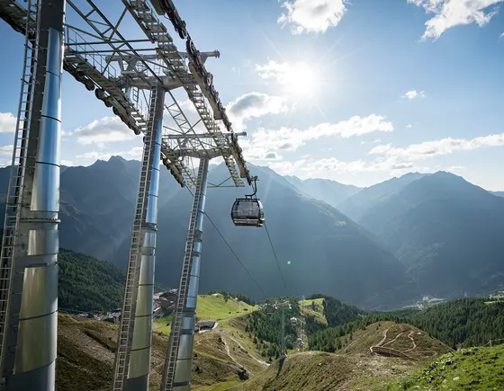 Ein Landschaftsfoto auf dem die Bergbahn und eine Gondel zu sehen sind. Grüne Wiesen, strahlend blauer Himmel mit ein paar Wolken über den Bergen.