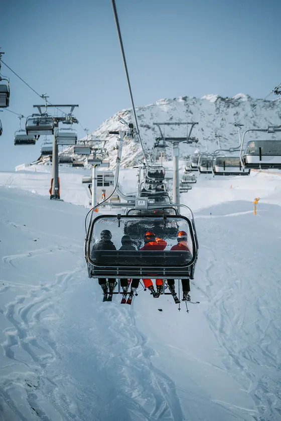 In der Mitte des Bildes sieht man vier Skifahrer auf einem Skilift sitzen, während sie mit dem Sessellift den Berg hinauffahren. Im Hintergrund sieht man die Skipisten.