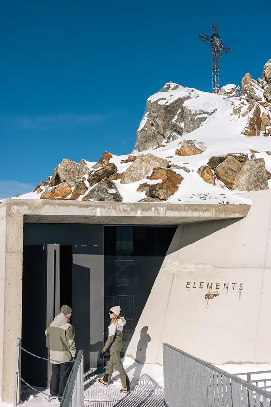 Zwei Personen stehen vor einem modernen Betongebäude in einer verschneiten, felsigen Berglandschaft unter einem strahlend blauen Himmel. Eingang vom Ausflugsziel 007 Elements im Ötztal.