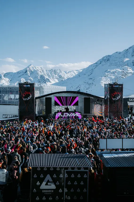 Eine große Menschenmenge versammelt sich bei einem Konzert im Freien mit verschneiten Bergen im Hintergrund unter einem klaren blauen Himmel.