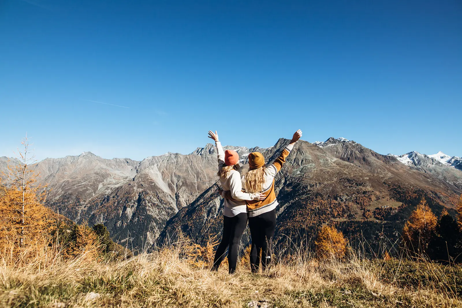 Zwei Menschen steht mit erhobenen Armen auf einer Bergwiese, mit malerischen Bergen und blauem Himmel im Hintergrund.