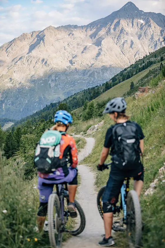 Zwei Radfahrerinnen tragen einen Helm und einen Rucksack, während sie auf einem Radweg stehen. Man sieht sie nur von hinten, links und rechts vom Radweg blühen grüne Wiesen und die Berge im Hintergrund. Sie machen auf einem Weg eine Pause mit Blick auf schroffe Berge und einen teilweise bewölkten Himmel.