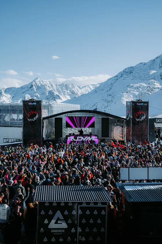 Die Bühne des Electric Mountain Festivals am Giggijoch mit den weißen Gipfelspitzen im Hintergrund und eine feiernde Menschenmenge davor.