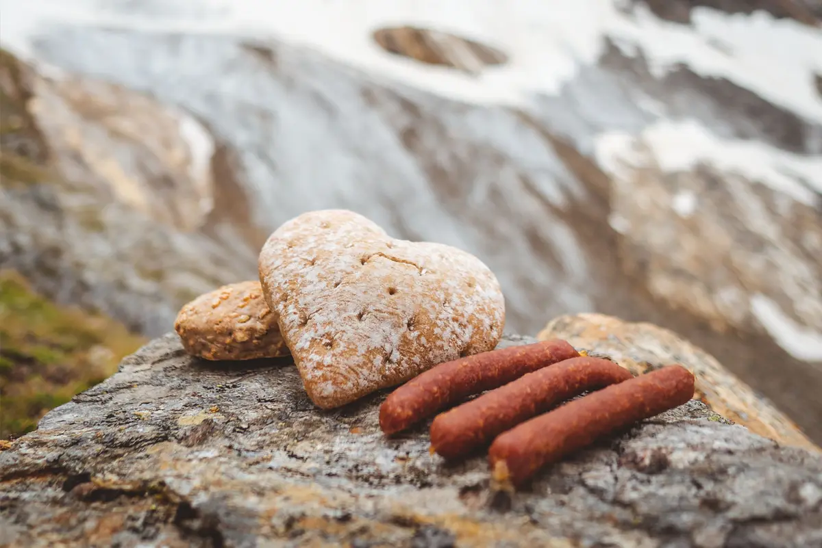 Auf einem Stein liegen drei Würstchen und daneben ein herzförmiges Brot mit Berghintergrund.