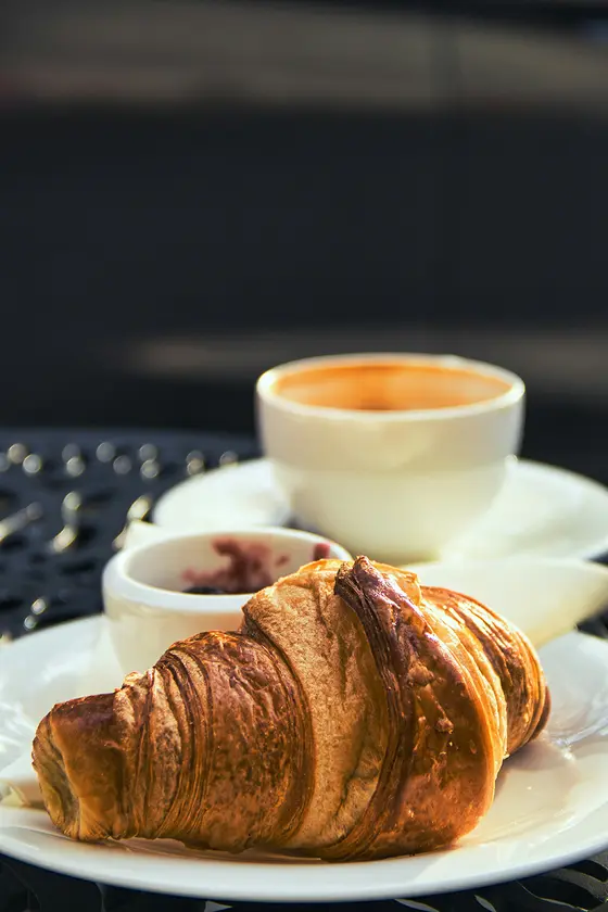 Ein Croissant auf einem Teller mit einer Tasse Kaffee und einer kleinen Schale mit Marmelade auf einem dunklen Tisch.