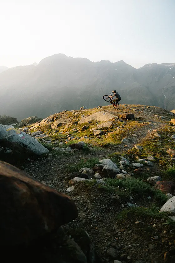 Eine Person sitzt auf einem Mountainbike auf einer grasbewachsenen Hügelkuppe mit Bergen im Hintergrund unter einem dunstigen Himmel.