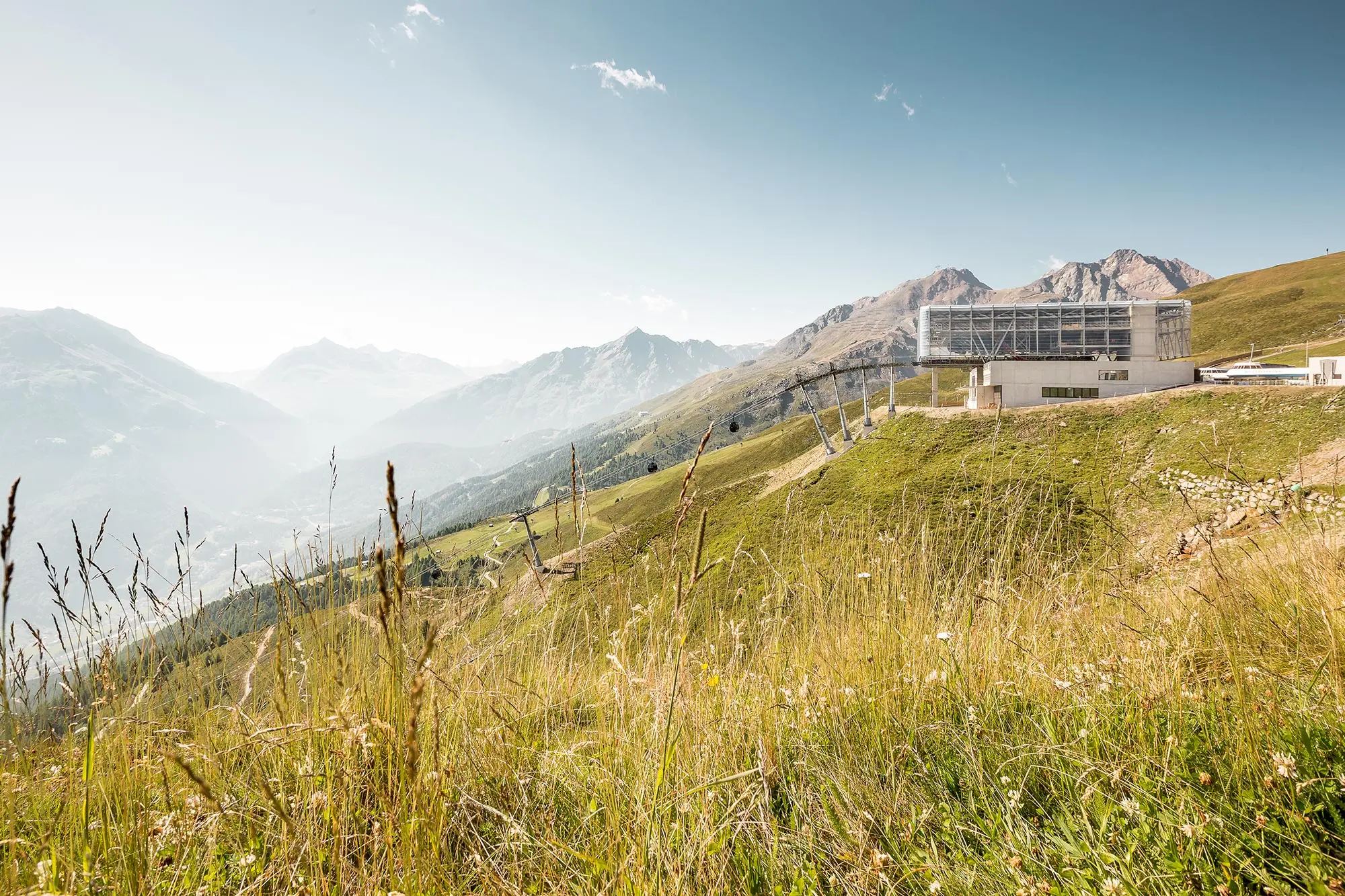 Eine Bergstation liegt auf einem grasbewachsenen Hügel mit weit entfernten schneebedeckten Gipfeln unter einem klaren blauen Himmel.