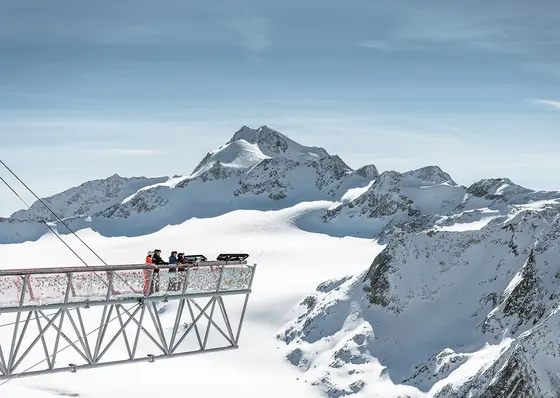 Menschen stehen auf einer metallenen Aussichtsplattform über verschneiten Bergen unter einem strahlend blauen Himmel mit Sonne und Wolken.