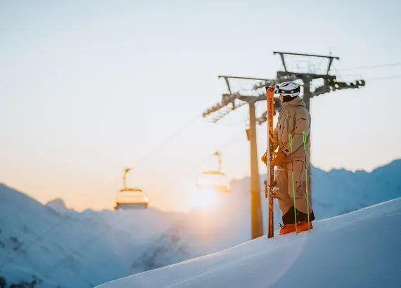 Skilifttürme und -seile bei Sonnenaufgang mit verschneiten Bergen im Hintergrund.