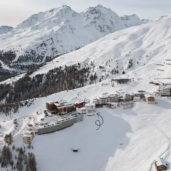Gebäude in einem Skigebiet, eingebettet in verschneite Berge mit Skifahrern und Sesselliften auf den Pisten. Schwarzer Pfeil zeigt auf den Standort der VIVO Hochsölden Apartments.