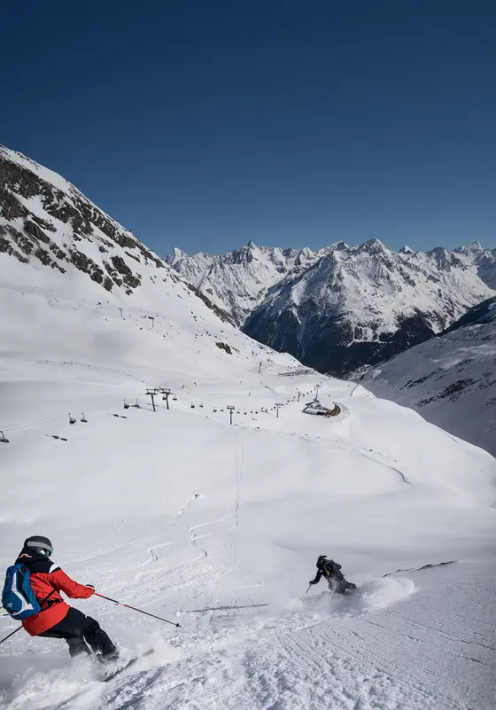 Ein Skifahrer in roter Jacke fährt durch Pulver-Schnee, umgeben von weißen Gipfeln und blauem Himmel.