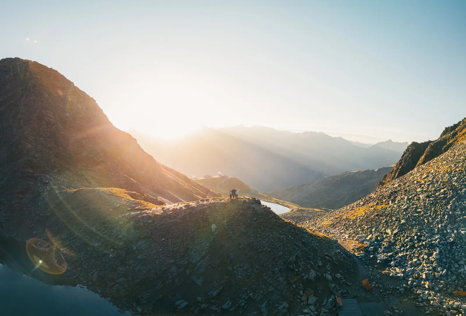 Links im Bild sieht man die Sonne, die sich hinter den Bergen versteckt. Sommerliche Berglandschaft mit grünen Wiesen.