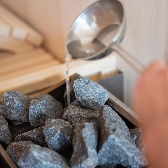 Eine Hand gießt Wasser aus einer Schöpfkelle auf heiße Saunasteine, wodurch Dampf entsteht.