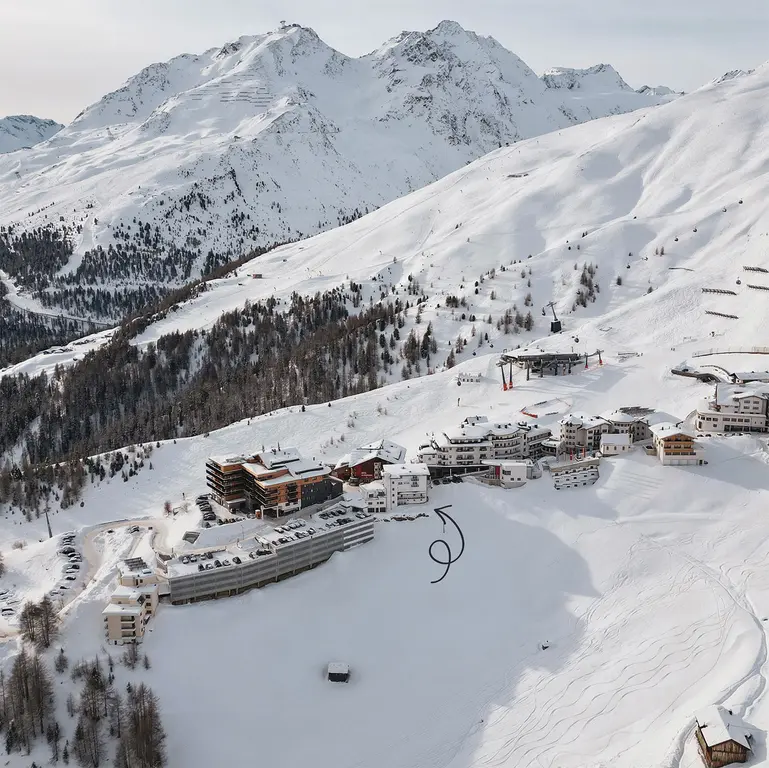 Vier Personen beim Skifahren auf einer verschneiten Bergpiste unter einem strahlenden Himmel, mit schneebedeckten Gipfeln im Hintergrund.