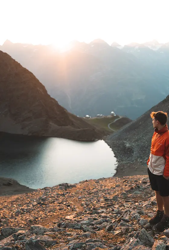 Eine Person in Outdoor-Ausrüstung steht auf einem felsigen Gelände mit Blick auf einen See und Berge bei Sonnenaufgang oder Sonnenuntergang.